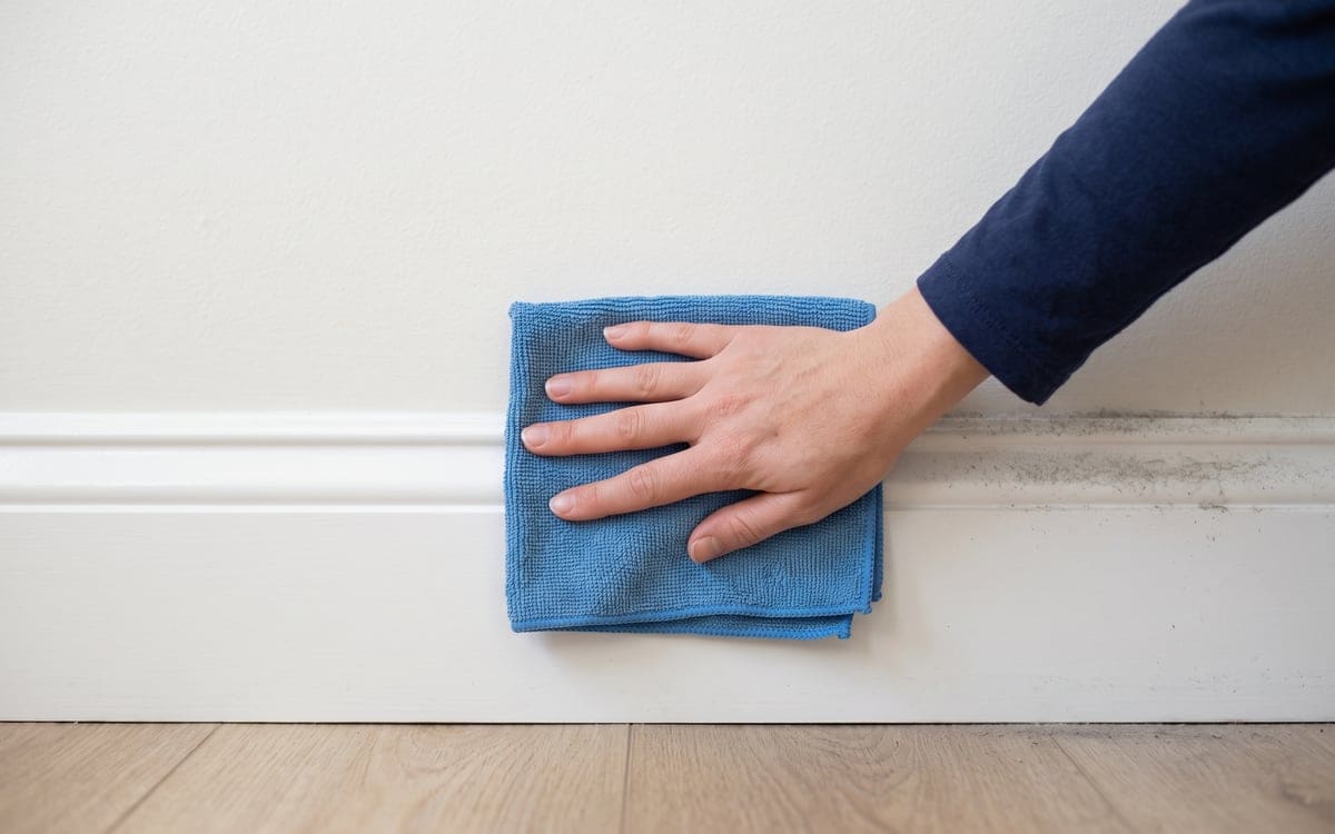 Along a white baseboard, a bare hand runs a folded blue microfiber cloth during a deep cleaning service, the wiped stretch bright and the stretch ahead still gray with dust.