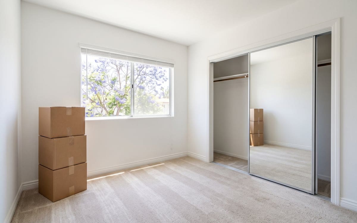 Three taped boxes stacked by the window of an empty San Diego bedroom on move-out day, a jacaranda in bloom outside and the mirrored closet slid open on a bare rod.