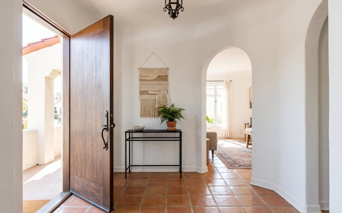 The tiled entry hall of a Spanish revival house after cleaning services, the wood front door open to the porch and an arched doorway beyond an iron console and a potted fern.