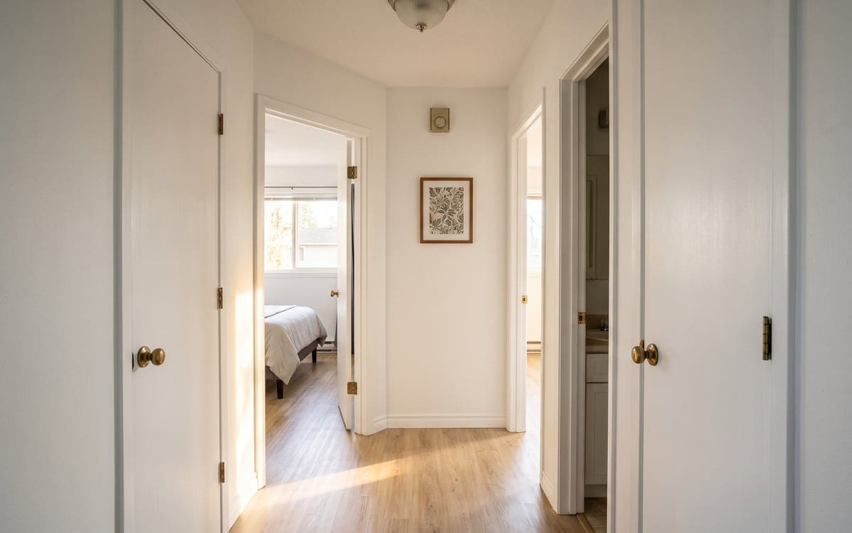 The short hallway of a two-bedroom condo, the bedroom and bathroom doors open, brass knobs on the closets and sun across the light plank floor.